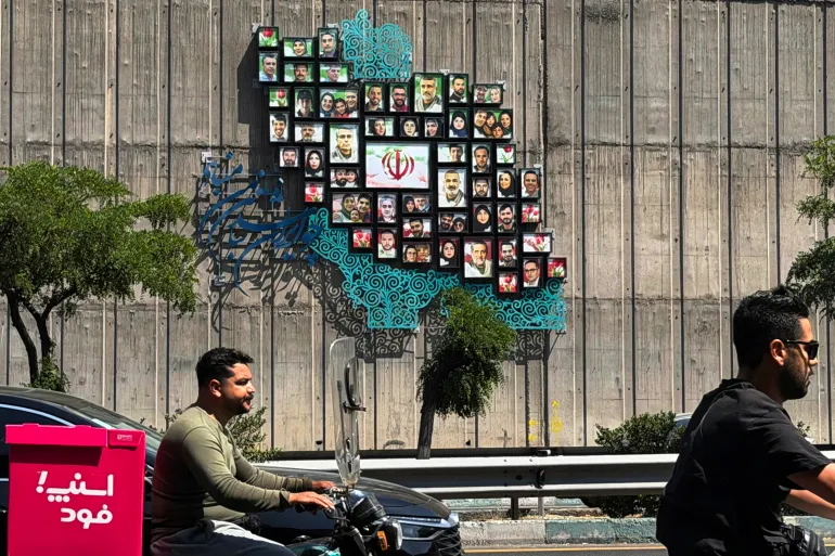 Hombres pasan en sus ciclomotores junto a un mapa de Irán con imágenes de algunos de los muertos en la guerra entre Israel y Estados Unidos contra la nación, erigido en un muro a lo largo de la carretera de Teherán el 17 de junio de 2026.